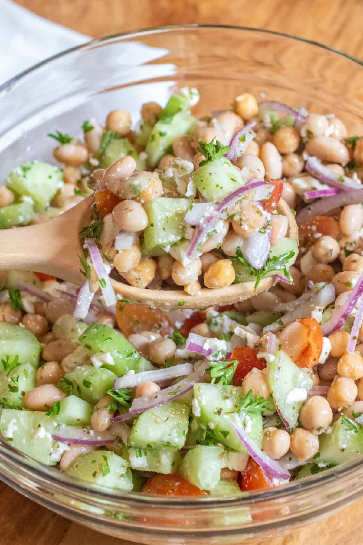 a bowl of chickpeas, beans, cucumber, tomatoes, onions, and feta with a spoonful in a wooden spoon.