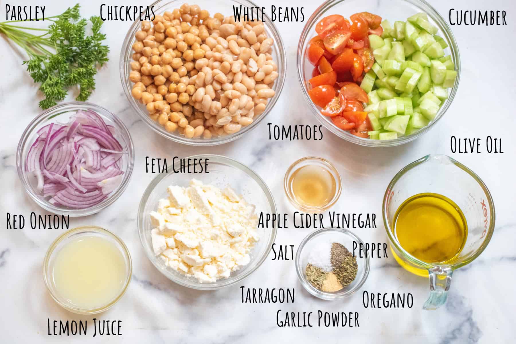 bowls of chickpeas, white beans, sliced onion, chopped tomatoes, cucumbers, feta, olive oil, lemon juice, apple cider vinegar, and salt, pepper, tarragon, oregano, and garlic powder all labeled with some fresh parsley on the side.