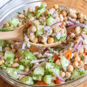 a bowl of chickpeas, beans, cucumber, tomatoes, onions, and feta with a spoonful in a wooden spoon.