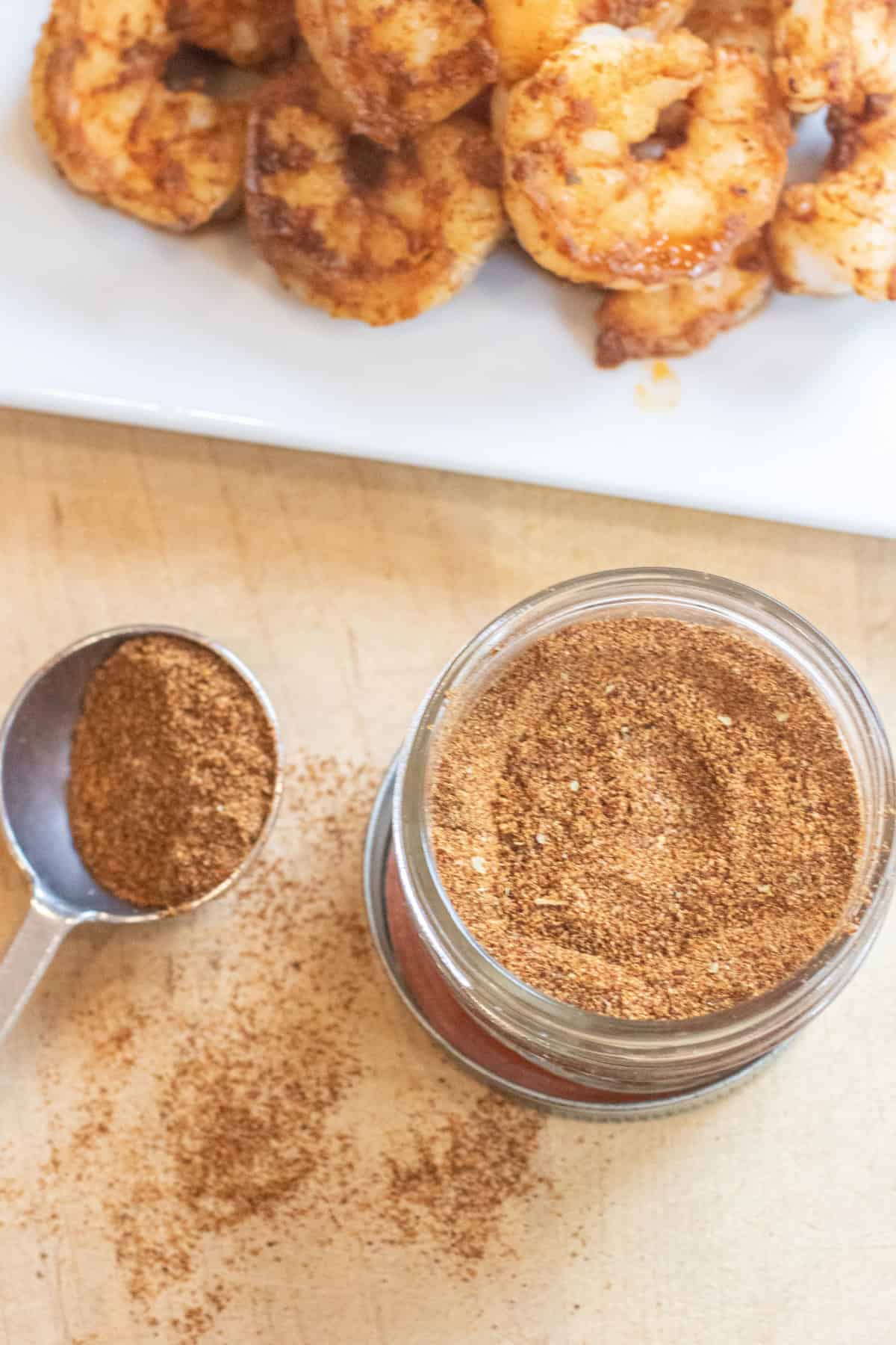 an overhead of a small mason jar of a spice blend mix with a measuring spoon next to it an some split in front of a plate of cooked shrimp.