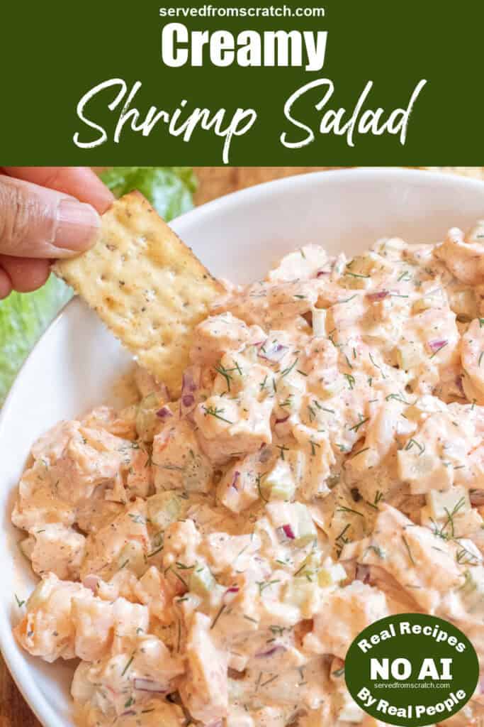an overhead of a big bowl of creamy shrimp salad with a hand dipping in a cracker with Pinterest pin text and NO AI sticker.