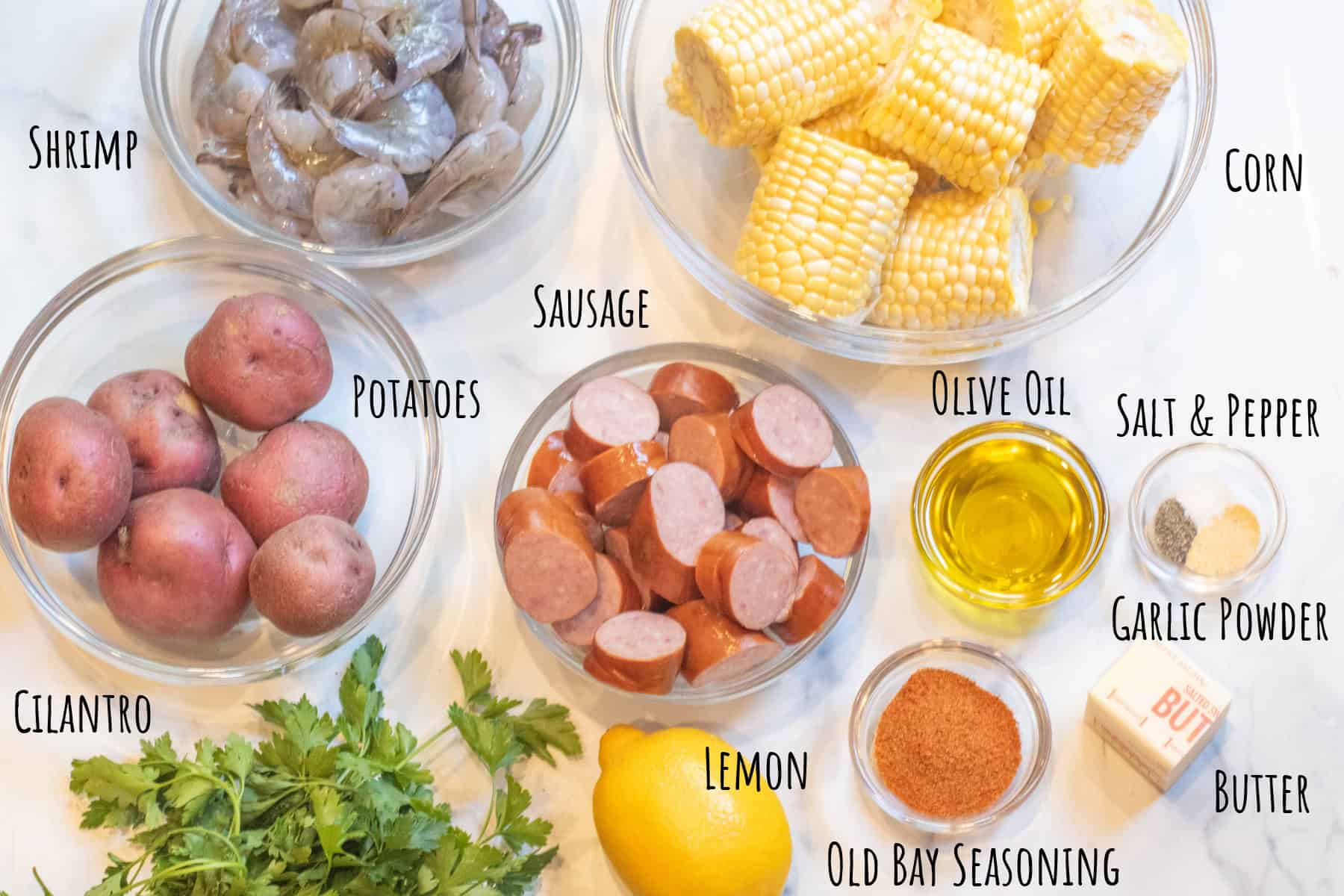 bowls of raw shrimp, corn, potatoes, sausage, oil, salt and pepper and garlic powder, old bay seasoning, butter, lemon, and parsley all labeled.