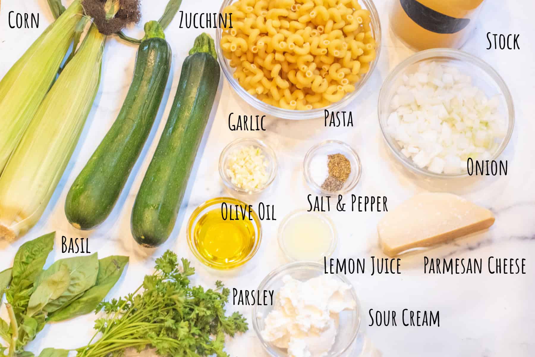 fresh zucchini, corn on the cob, a bowl of pasta, cheeses, chicken stock, spices, olive oil, fresh herbs, and parmesan cheese all on a counter and labeled.