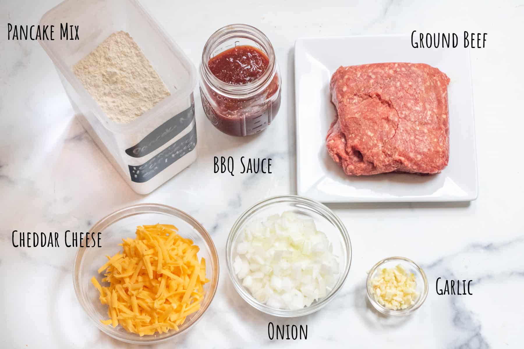 a container or pancake mix, an open mason jar of BBQ sauce, a plate with ground beef, a bowl of cheddar cheese and a bowl of diced onion and a bowl of minced garlic all labeled.