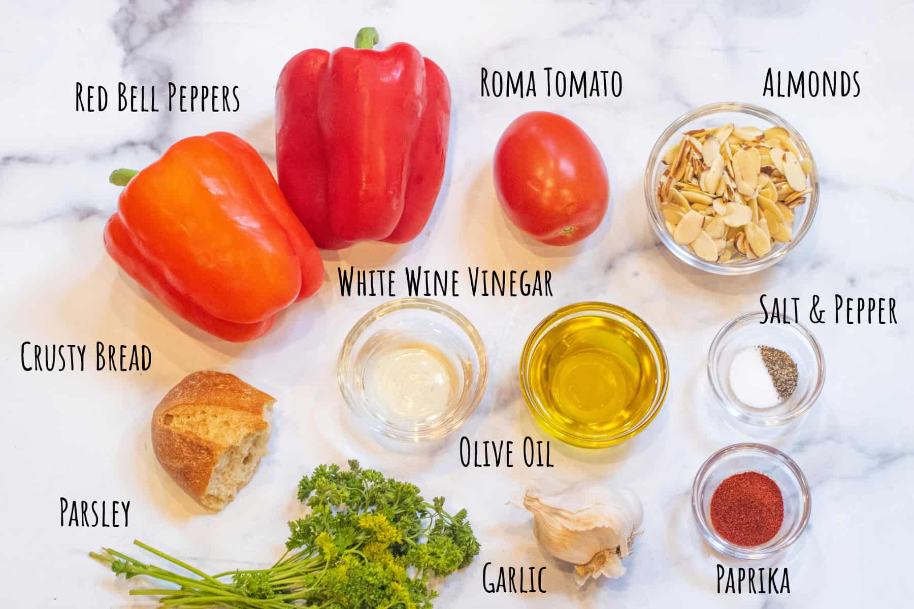 red peppers, a roma tomato, sliced almonds, vinegar, olive oil, salt and pepper, crusty bread, fresh parsley, garlic, and paprika labeled in bowls.