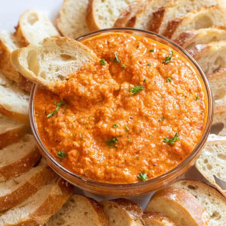 a bowl of romesco dip with a slice of baguette surrounded by baguette slices on a plate.