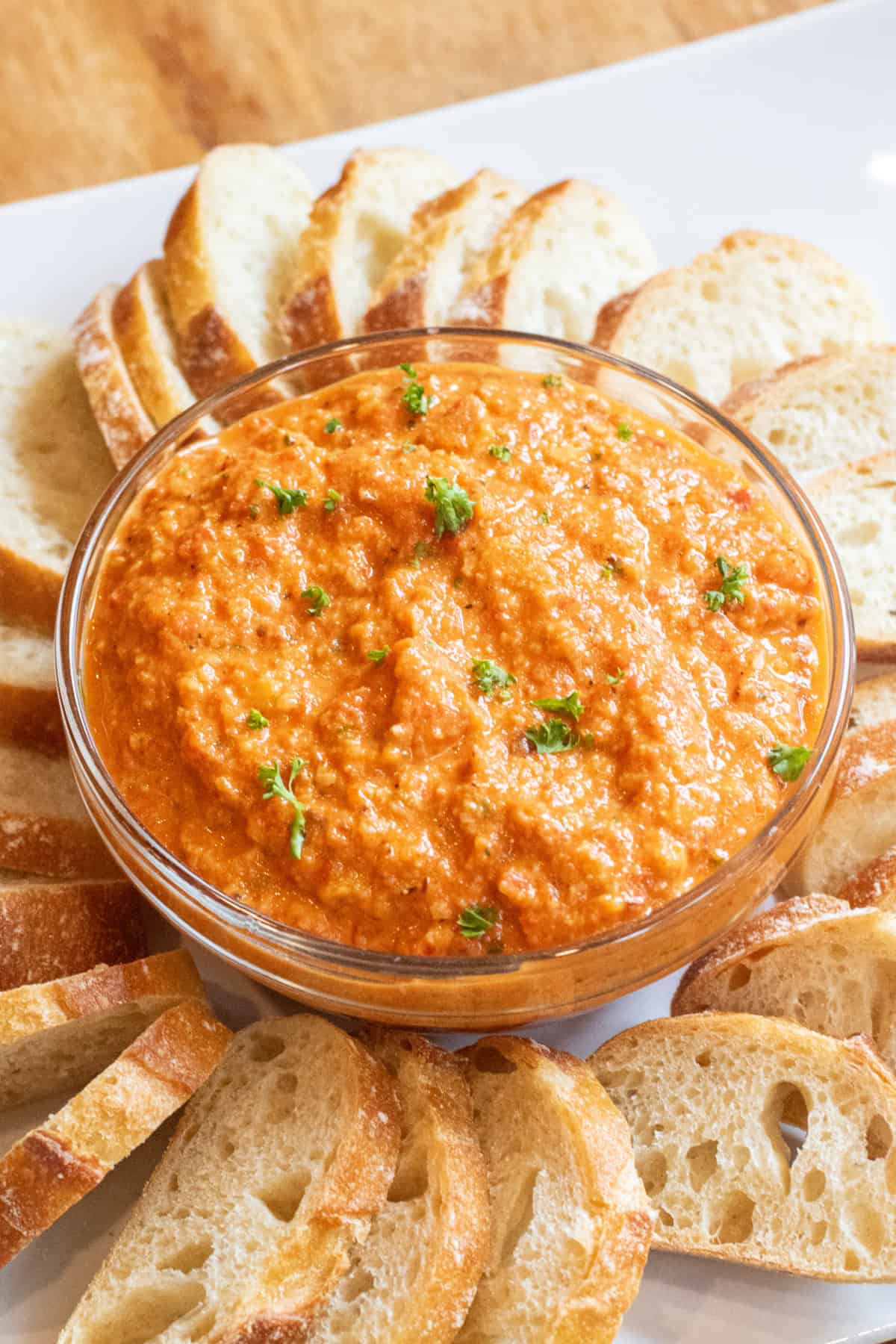 a bowl of romesco dip with a slice of baguette surrounded by baguette slices on a plate.
