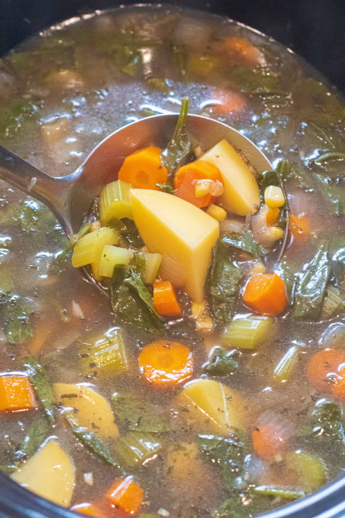an overhead of a ladle in a crock pot with soup of potatoes, carrots, corn, onions, spinach.