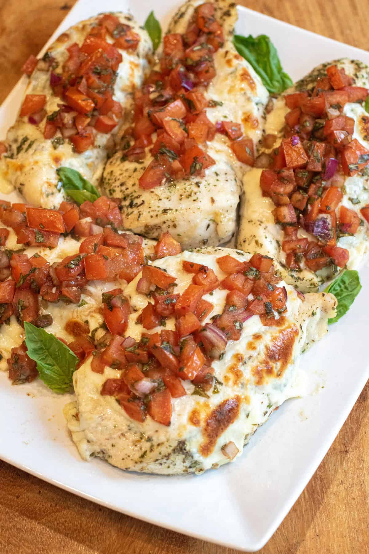 a plate of baked chicken breasts topped with melted cheese and tomato bruschetta.